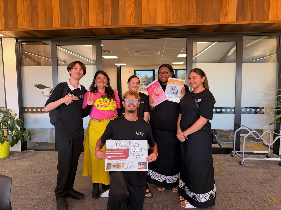 Group photo of Kaitaia College interns from Te Hiku Hauora taken in an office setting showing six people participating in a youth-focused project. One participant kneels in front holding a display board, while others stand behind holding posters and educational materials.