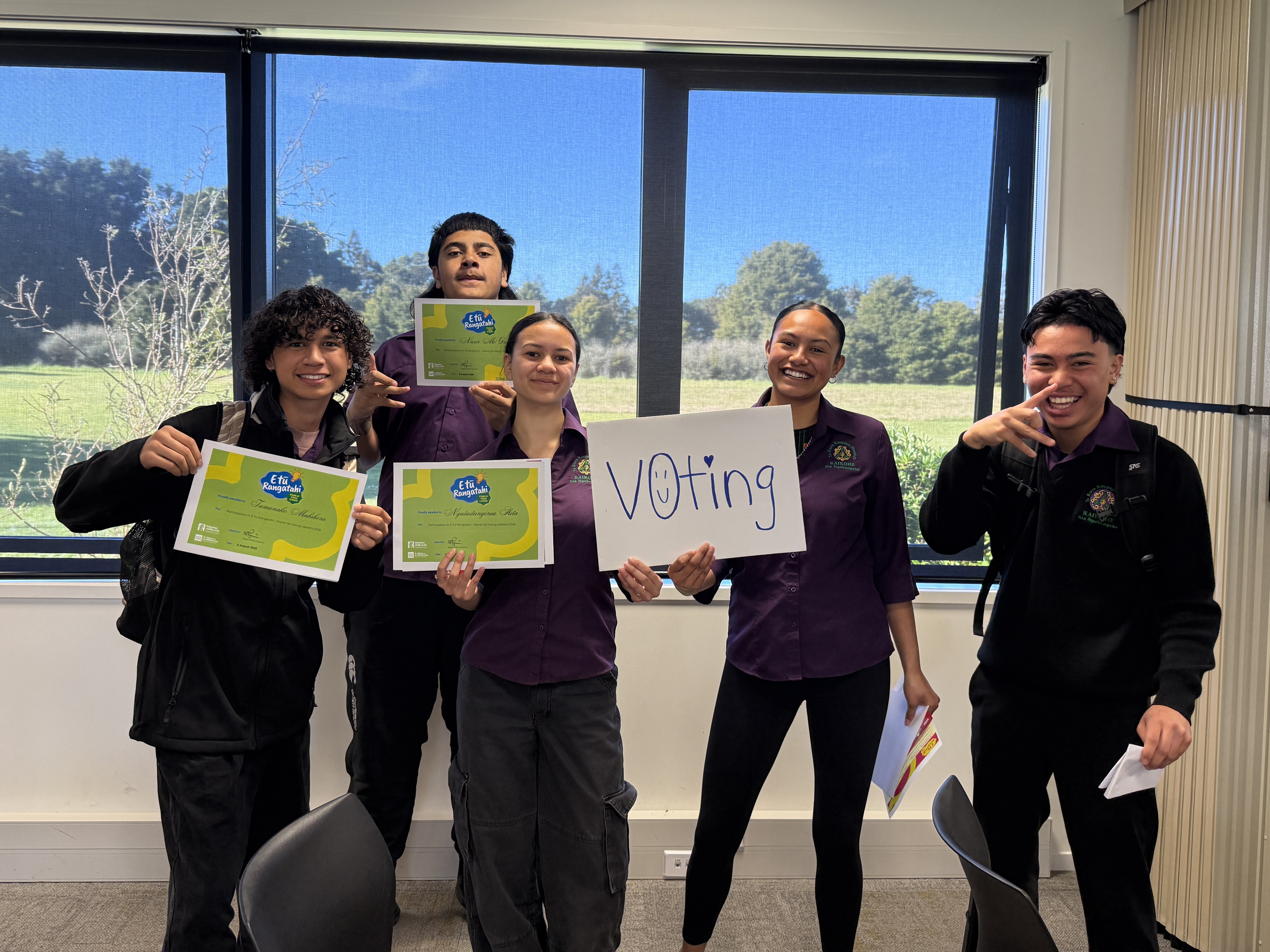 Five students from Te Kura Kaupapa Māori o Kaikohe pose with participation certificates and a handwritten &ldquo;voting&rdquo; sign in a classroom or meeting room. Large windows behind the group overlook a green outdoor landscape.